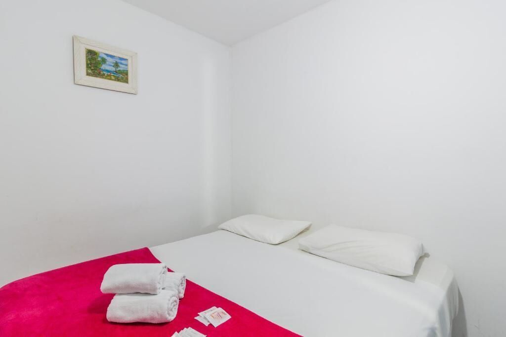 A single bed with white linens and pillows, a red blanket, and two rolled towels on the floor.