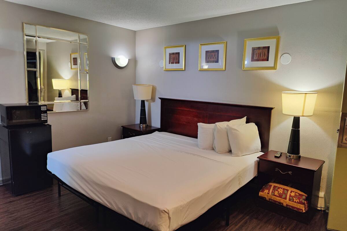 Une chambre d’hôtel avec un grand lit, une lampe de chevet, une table de chevet avec une télécommande, une petite table de chevet avec une lampe, un miroir et des tableaux encadrés au mur.