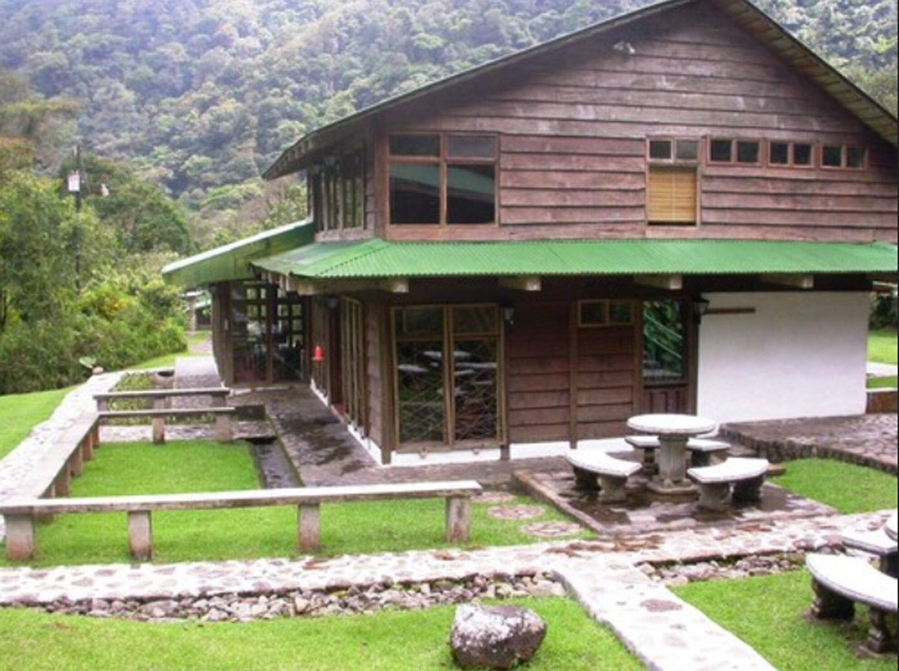 A rustic wooden building with a green roof, a patio area with stone benches, and a stone pathway leading to the entrance.