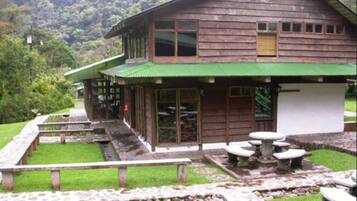 A rustic wooden building with a green roof, a patio area with stone benches, and a stone pathway leading to the entrance.