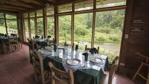 A dining area with tables set for four, large windows offering a view of a river and greenery, and a wooden ceiling structure.