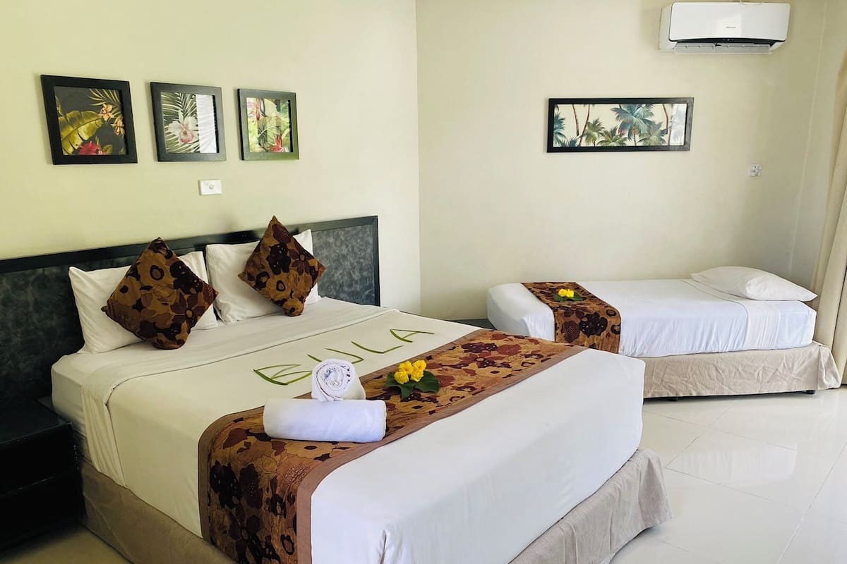 Une chambre d’hôtel avec deux lits : un grand lit recouvert d’une courtepointe à motifs floraux et agrémenté d’oreillers, et un plus petit lit recouvert d’une simple couverture. Trois tableaux encadrés ornent le mur, ainsi qu’un appareil de climatisation et une petite table sur laquelle reposent une serviette et des fleurs sur le grand lit.