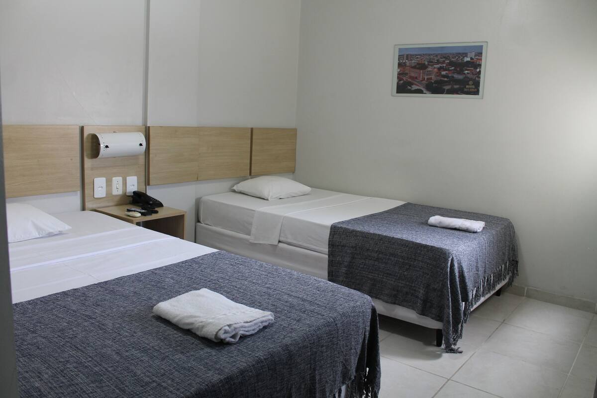 Une chambre d’hôtel avec deux lits simples, une tête de lit en bois, une table de chevet avec un téléphone et une lampe, et un distributeur de serviettes fixé au mur.