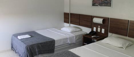 A hotel room with two single beds, a wooden headboard, a nightstand with a phone and remote, and a framed cityscape picture on the wall.
