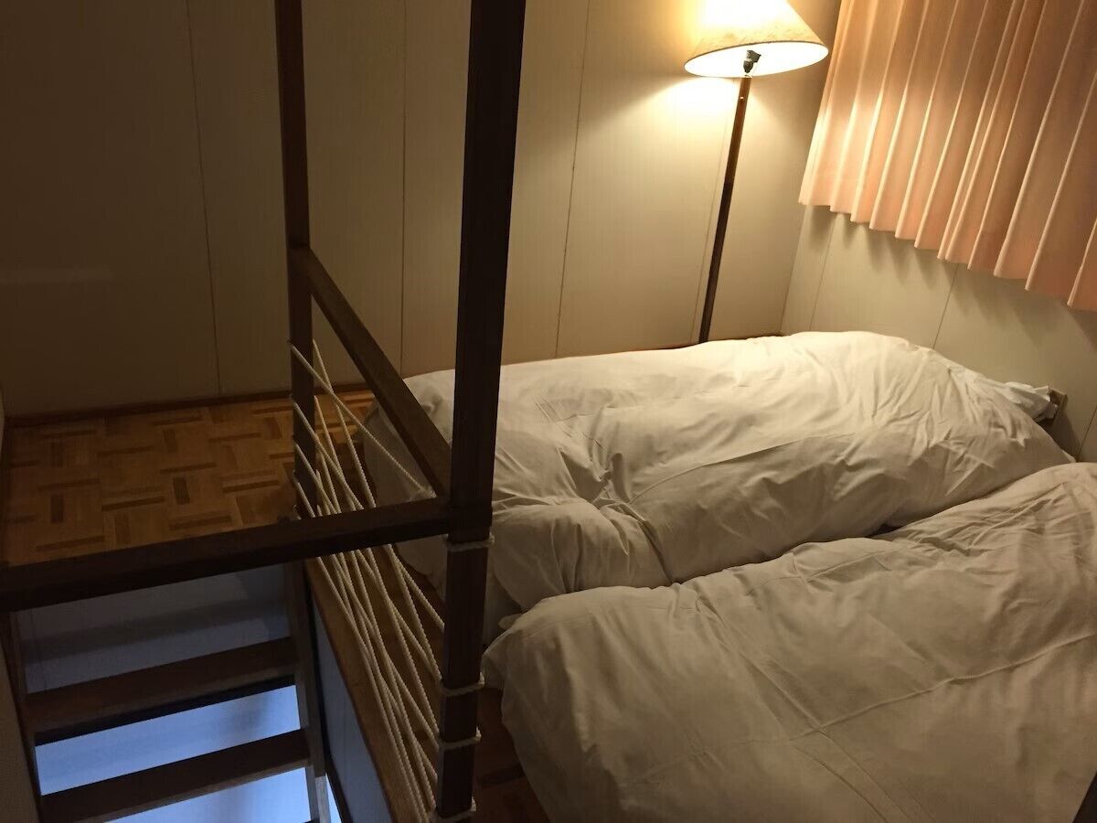 A bunk bed with a wooden ladder, a bedside lamp, and a window with blinds.