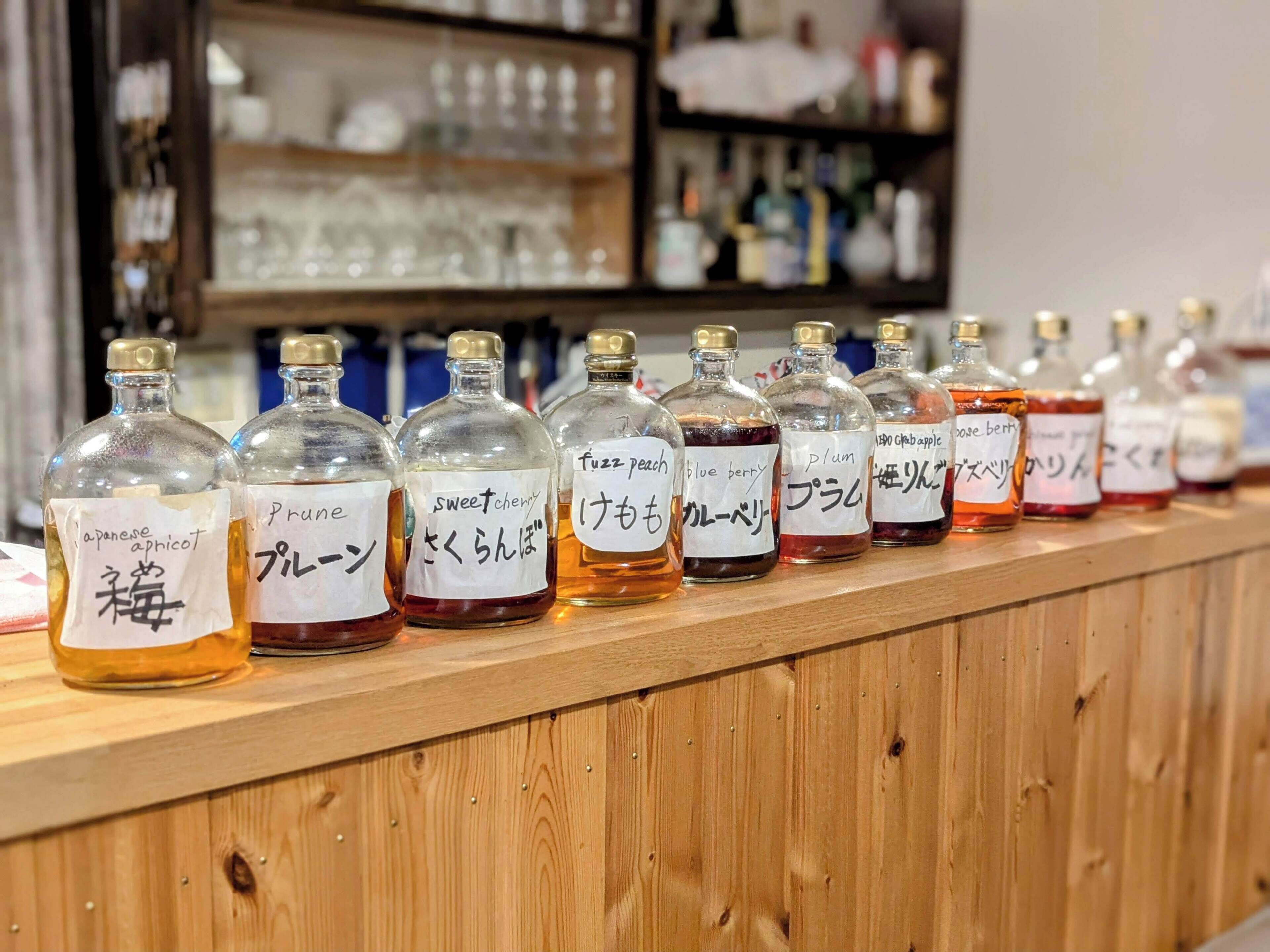 A row of glass bottles with labels in multiple languages, likely containing flavored syrups or similar products, displayed on a wooden counter.