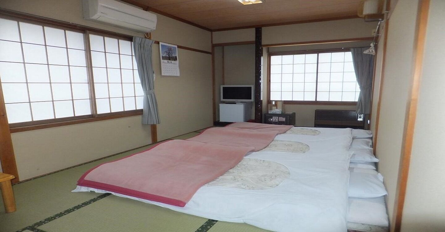 Une pièce de style japonais traditionnel, avec un sol recouvert de tatamis, un lit avec une couvre-lit rose et blanche, une télévision et une petite table.