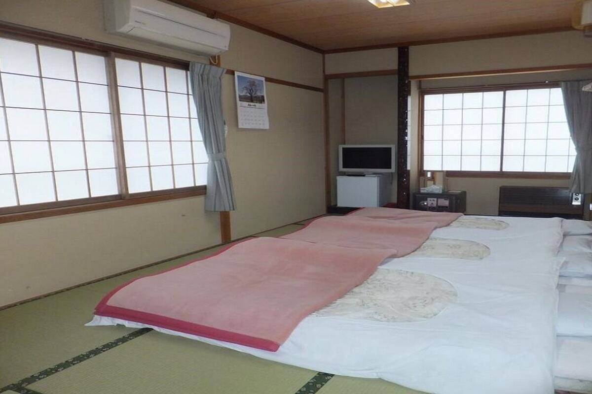 Une pièce de style japonais traditionnel, avec un sol recouvert de tatamis, un lit avec une couvre-lit rose et blanche, une télévision et une petite table.