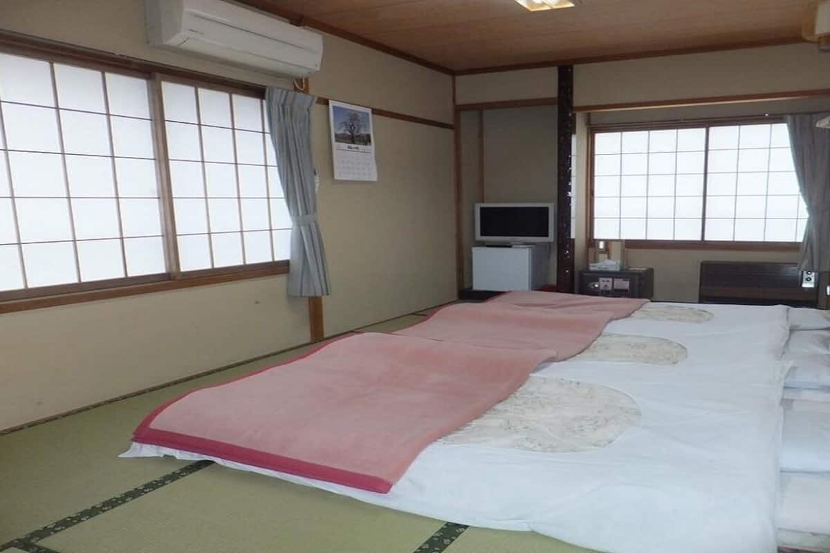 Une pièce de style japonais traditionnel, avec un sol recouvert de tatamis, un lit avec une couvre-lit rose et blanche, une télévision et une petite table.