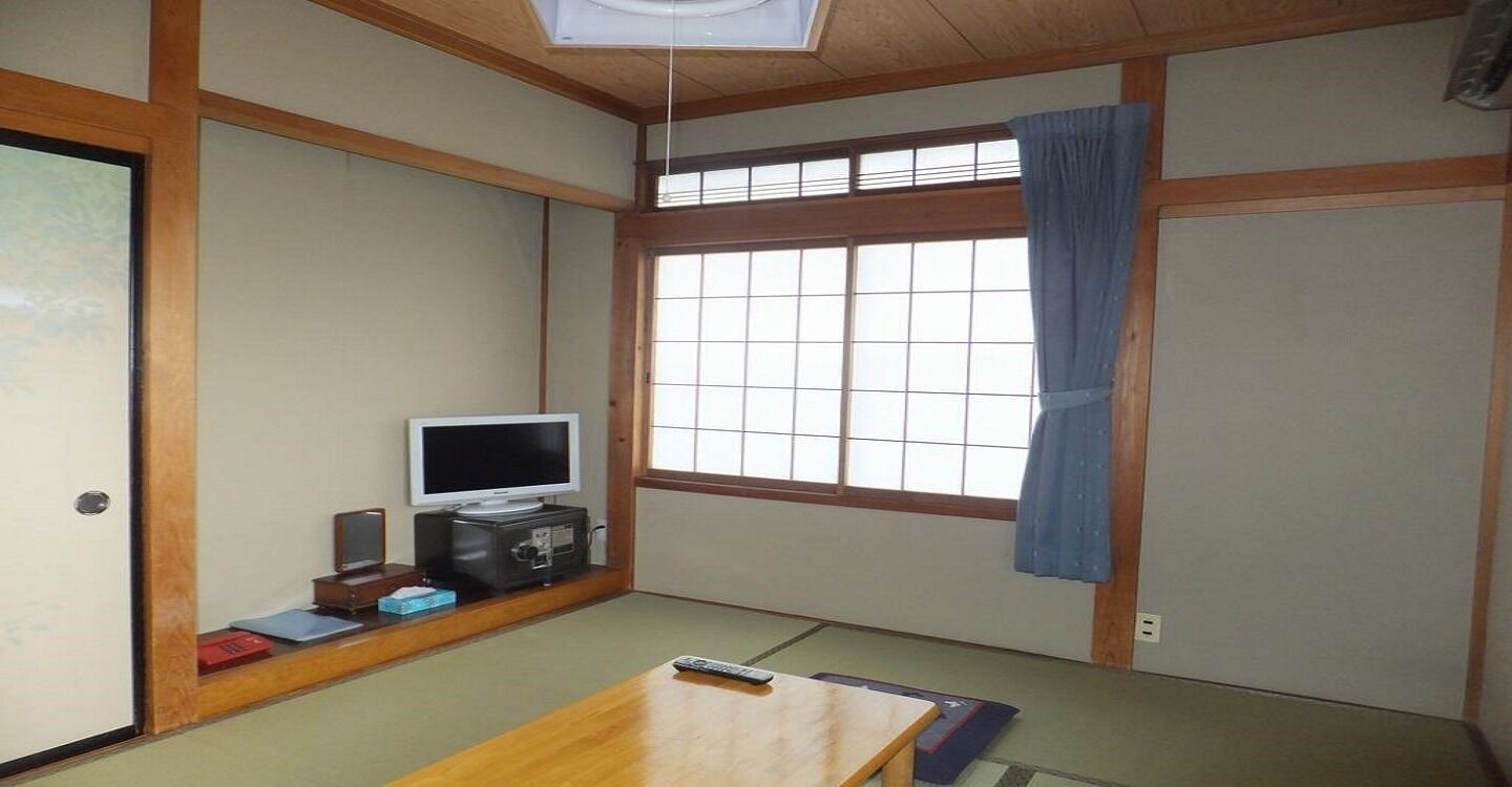 Une pièce de style japonais traditionnel, avec un sol recouvert de tatamis, un téléviseur, une petite armoire et une fenêtre avec un rideau.
