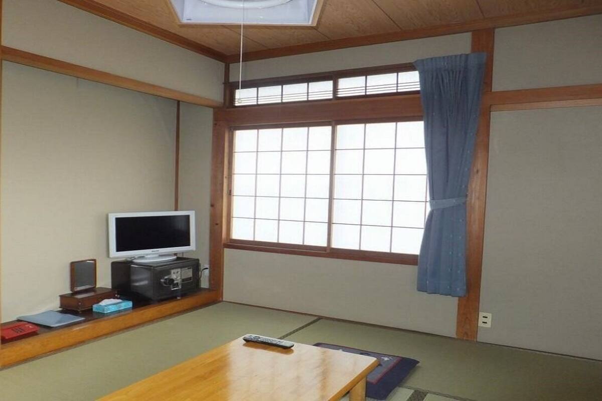 Une pièce de style japonais traditionnel, avec un sol recouvert de tatamis, un téléviseur, une petite armoire et une fenêtre avec un rideau.