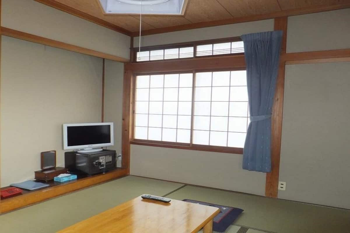 Une pièce de style japonais traditionnel, avec un sol recouvert de tatamis, un téléviseur, une petite armoire et une fenêtre avec un rideau.