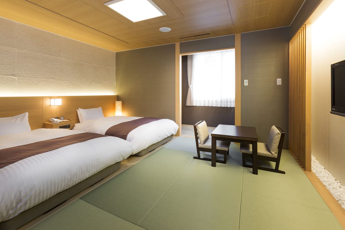 A hotel room with two beds, a small table with chairs, a TV, and a window with curtains.