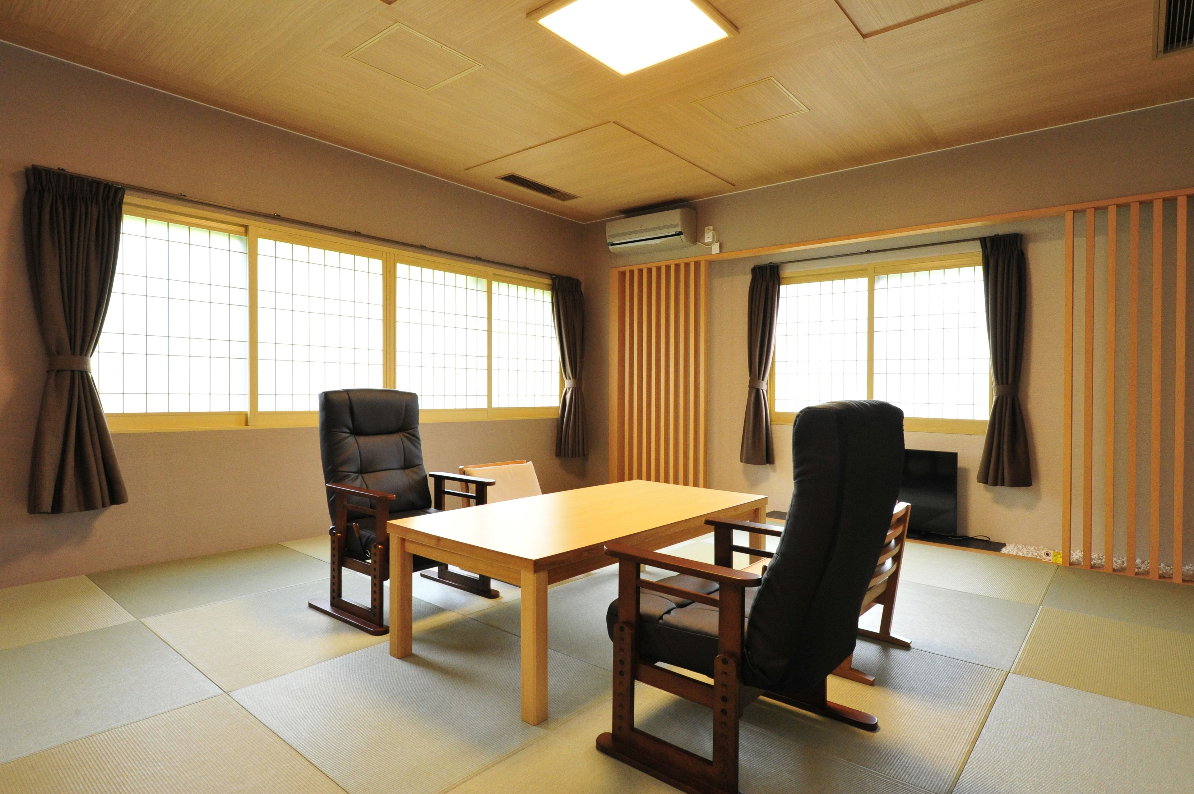 Ein traditioneller japanischer Raum mit Tatami-Bodenbelag, einem niedrigen Tisch, zwei Stühlen und großen Fenstern mit Vorhängen.