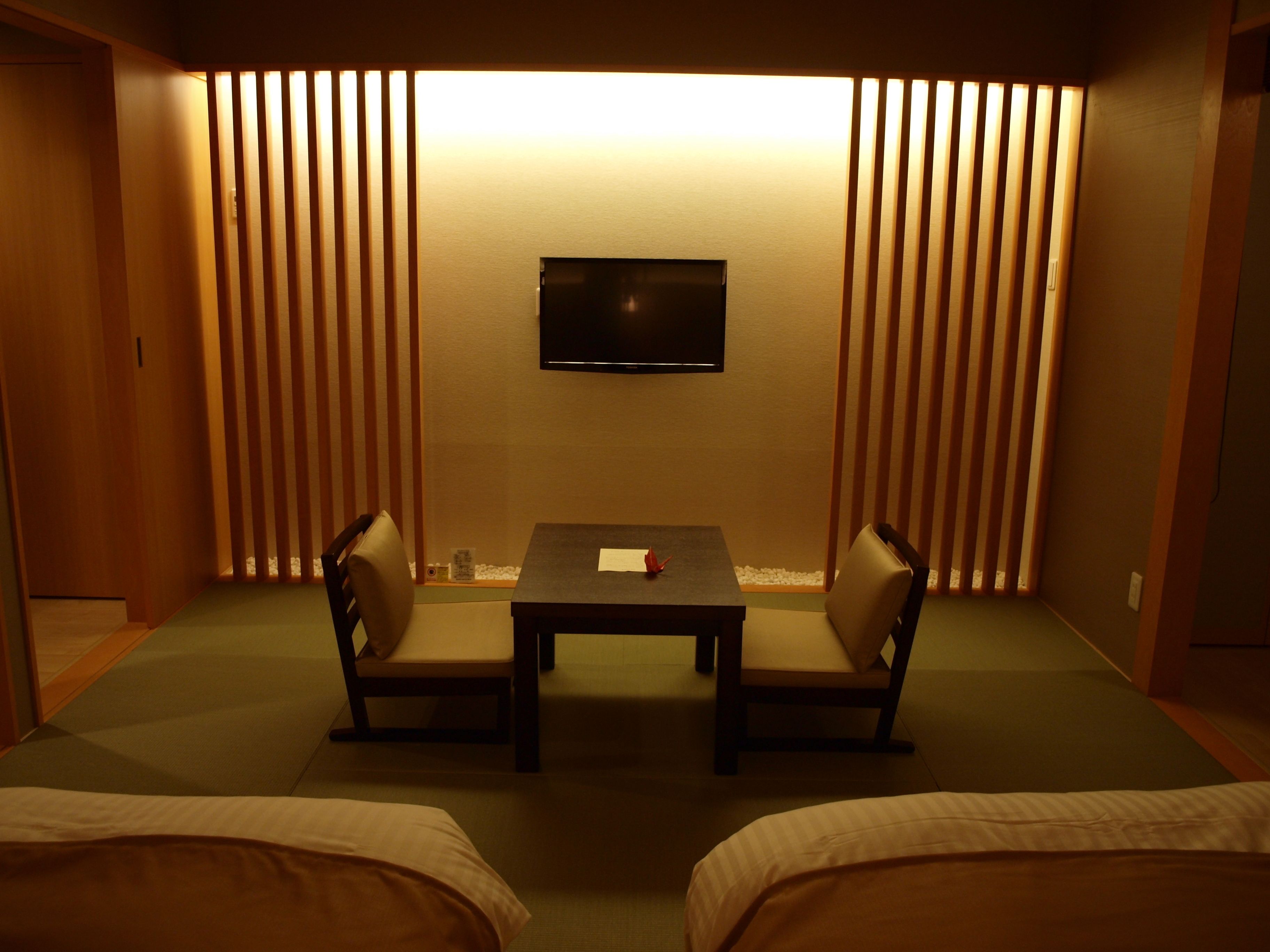 A room with a TV mounted on the wall, a small table with chairs, and a bed with a green mat.