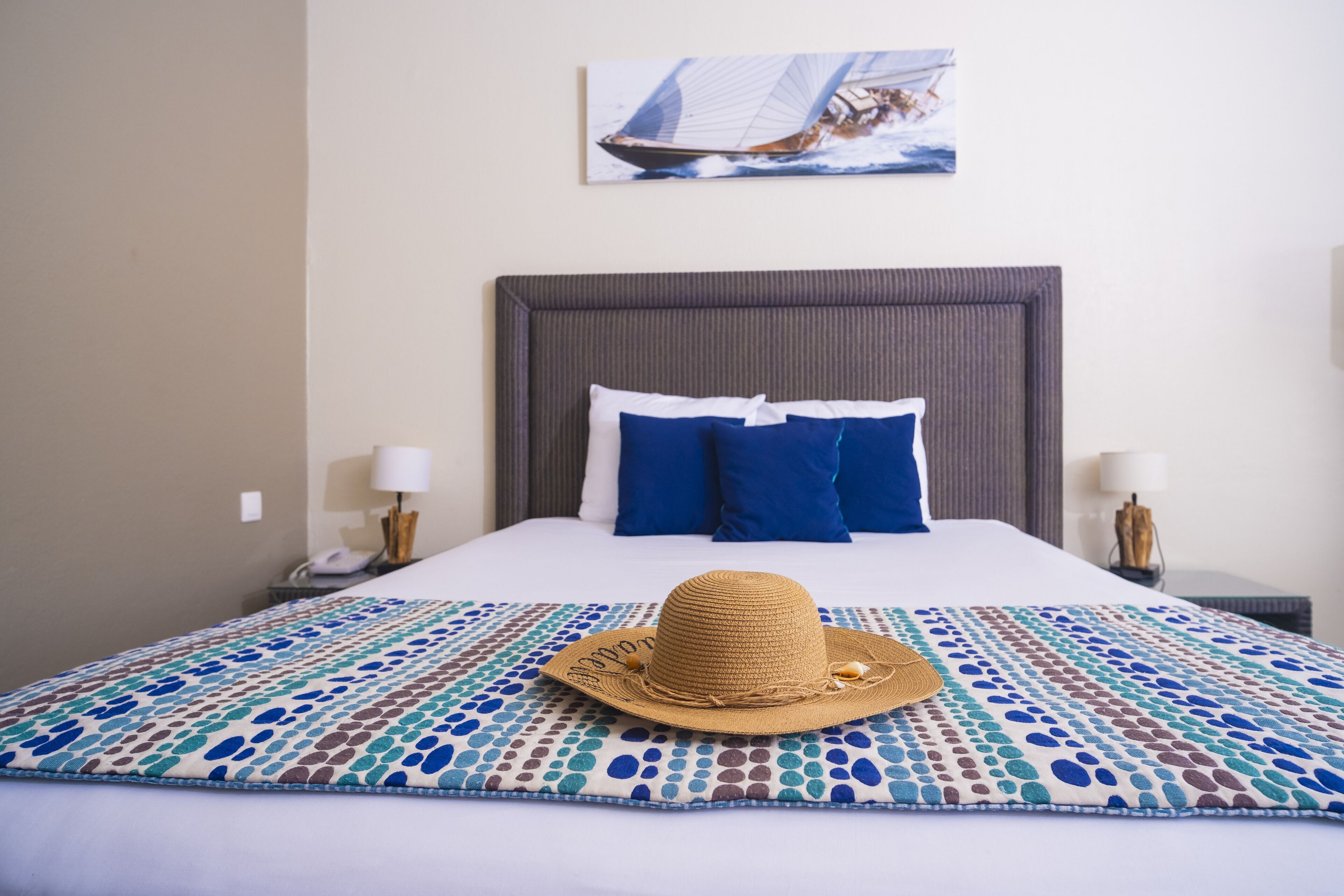 A neatly made bed with a patterned bedspread, a straw hat on the bed, and a sailboat painting on the wall.