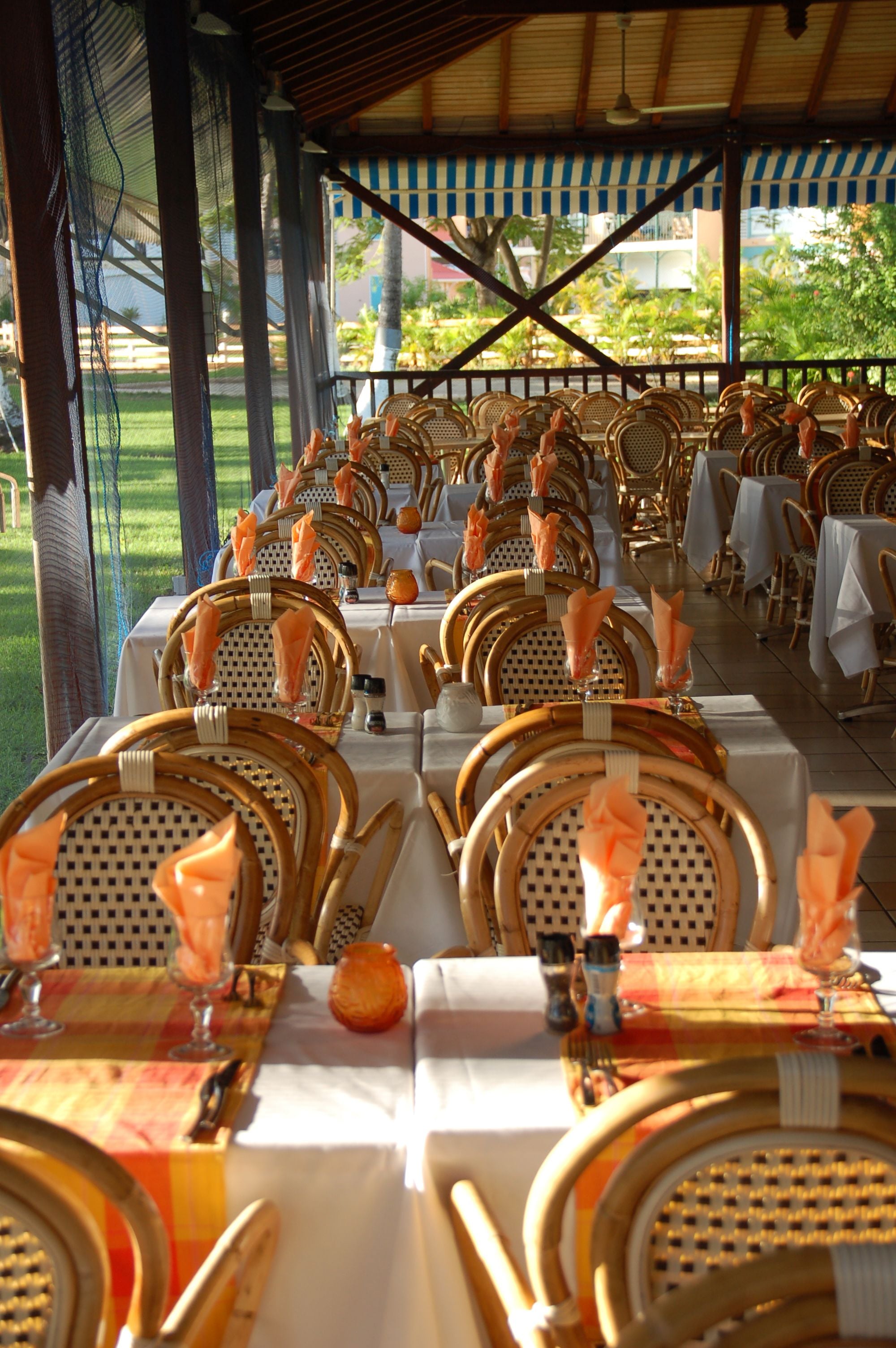 Een terras met eettafels gedekt voor meerdere gasten, met rieten stoelen en witte tafelkleden.