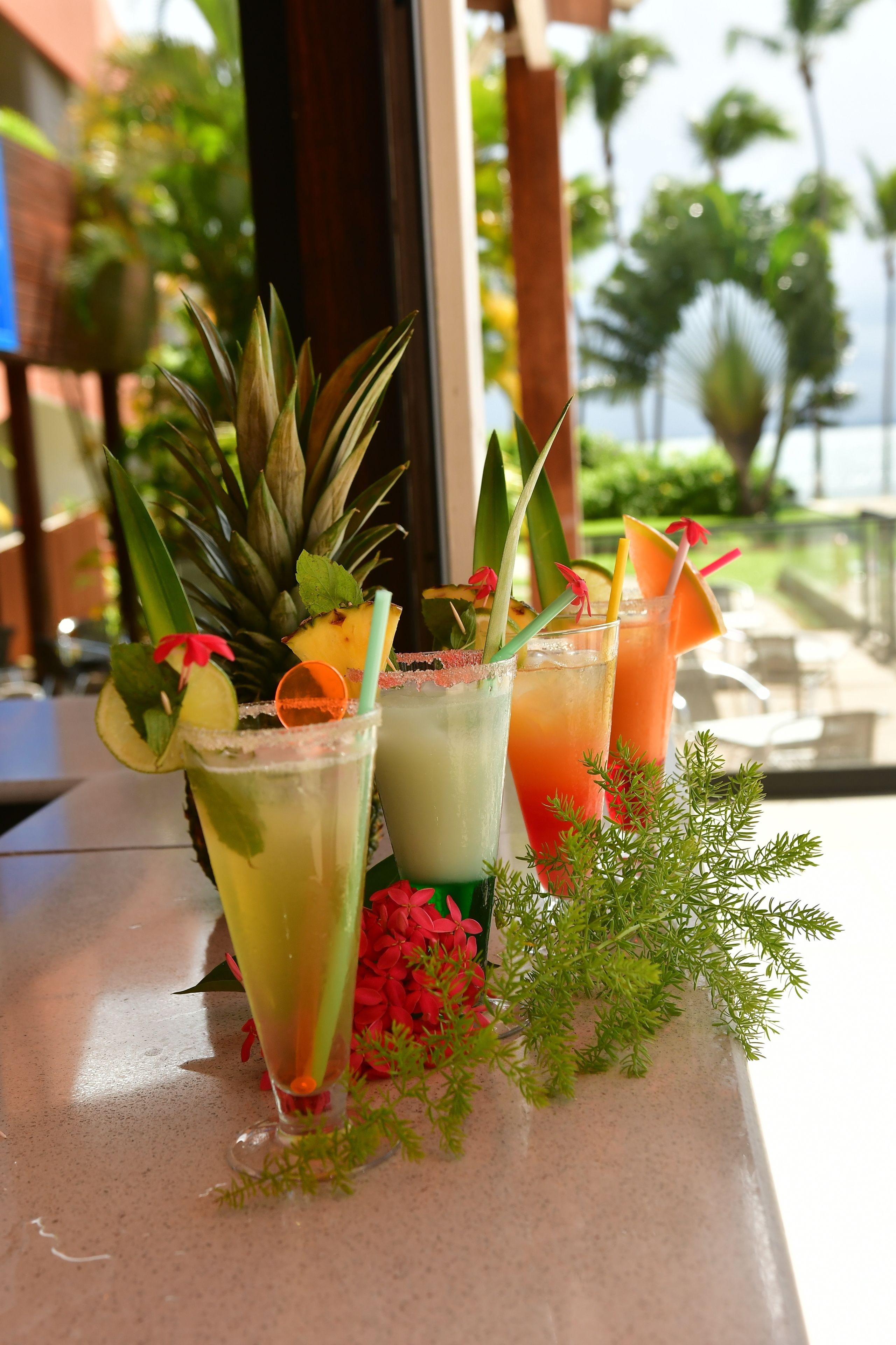 Tre eksotiske cocktails på et bord med udsigt til palmer og en bygning i baggrunden.