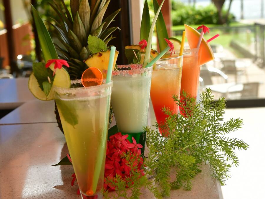 Três cocktails tropicais sobre uma mesa, com vista para palmeiras e um prédio ao fundo.