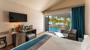 A hotel room with a bed, a desk with a flat-screen TV, a small table, and a chair. There is a balcony with a view of a pool and palm trees. - Hotel Fleur d'Epée (Le Gosier)