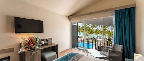 Habitación de hotel con cama, escritorio con televisor de pantalla plana, una mesita y una silla. Tiene balcón con vista a una piscina y palmeras.