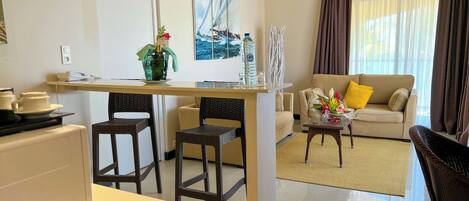Habitación de hotel con kitchenette, zona de comedor con sillas, sofá y ventana grande con cortinas.