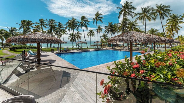 A pool area with thatched umbrellas, lounge chairs, and a view of the ocean. - Hotel Fleur d'Epée (Le Gosier)