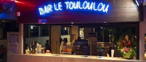 A bar named "BAR LE TOULOUAU" with a neon sign, a counter with a beverage dispenser, and a seating area with chairs.