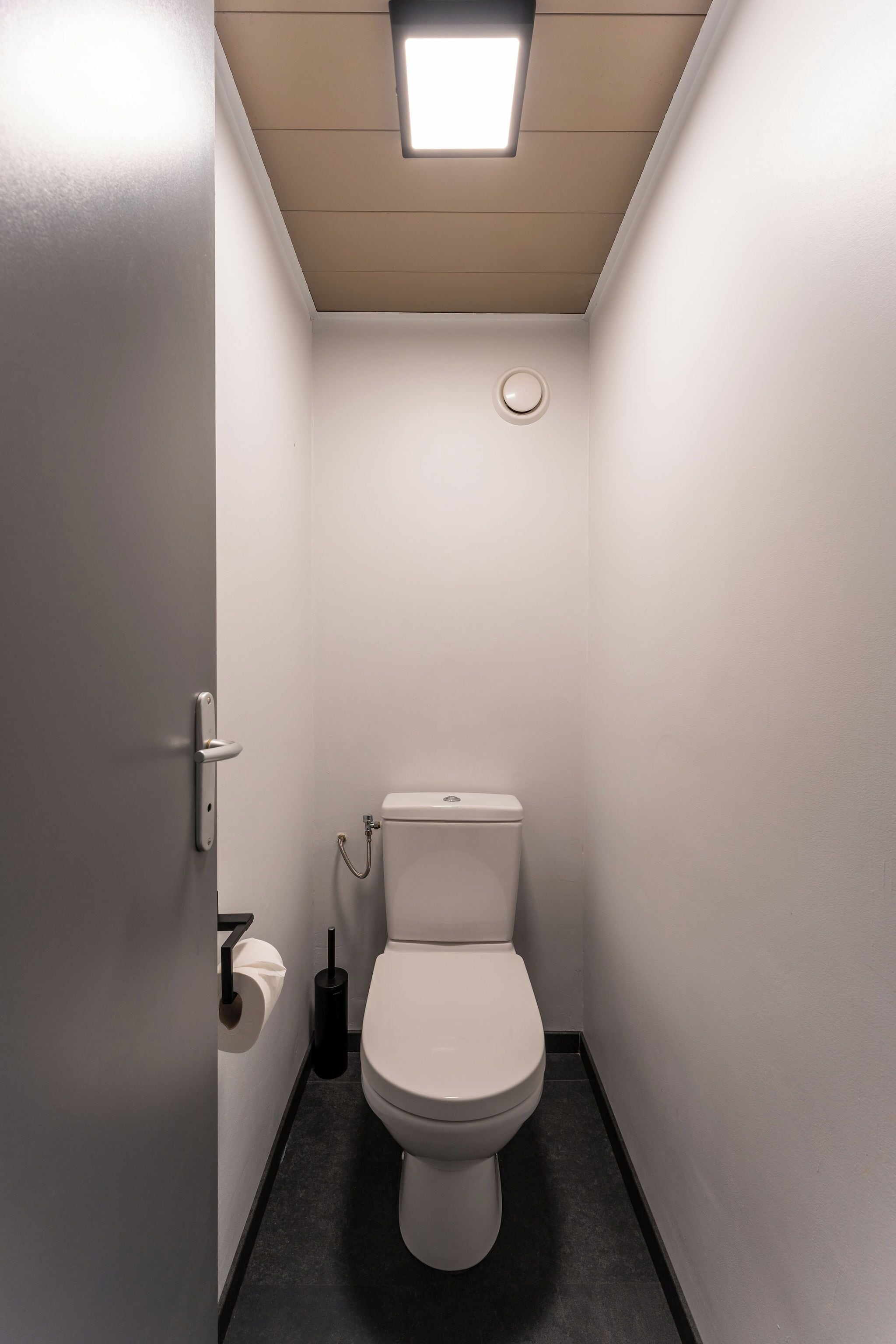 A small, narrow bathroom with a white toilet, a toilet paper holder, and a flush handle.