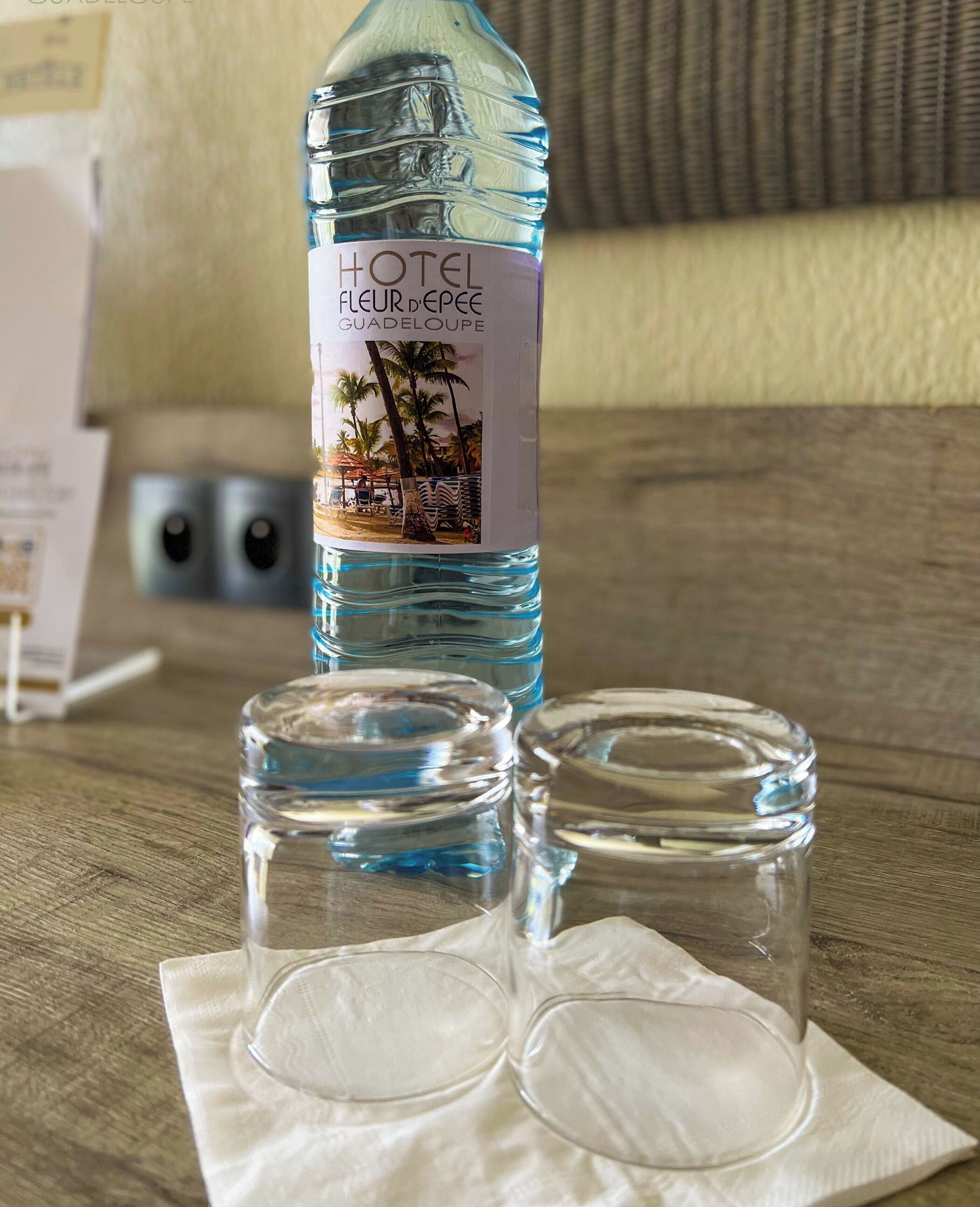 Una botella de agua Hotel Fleur de Epee Guadeloupe, dos vasos y una servilleta sobre una mesa de madera.