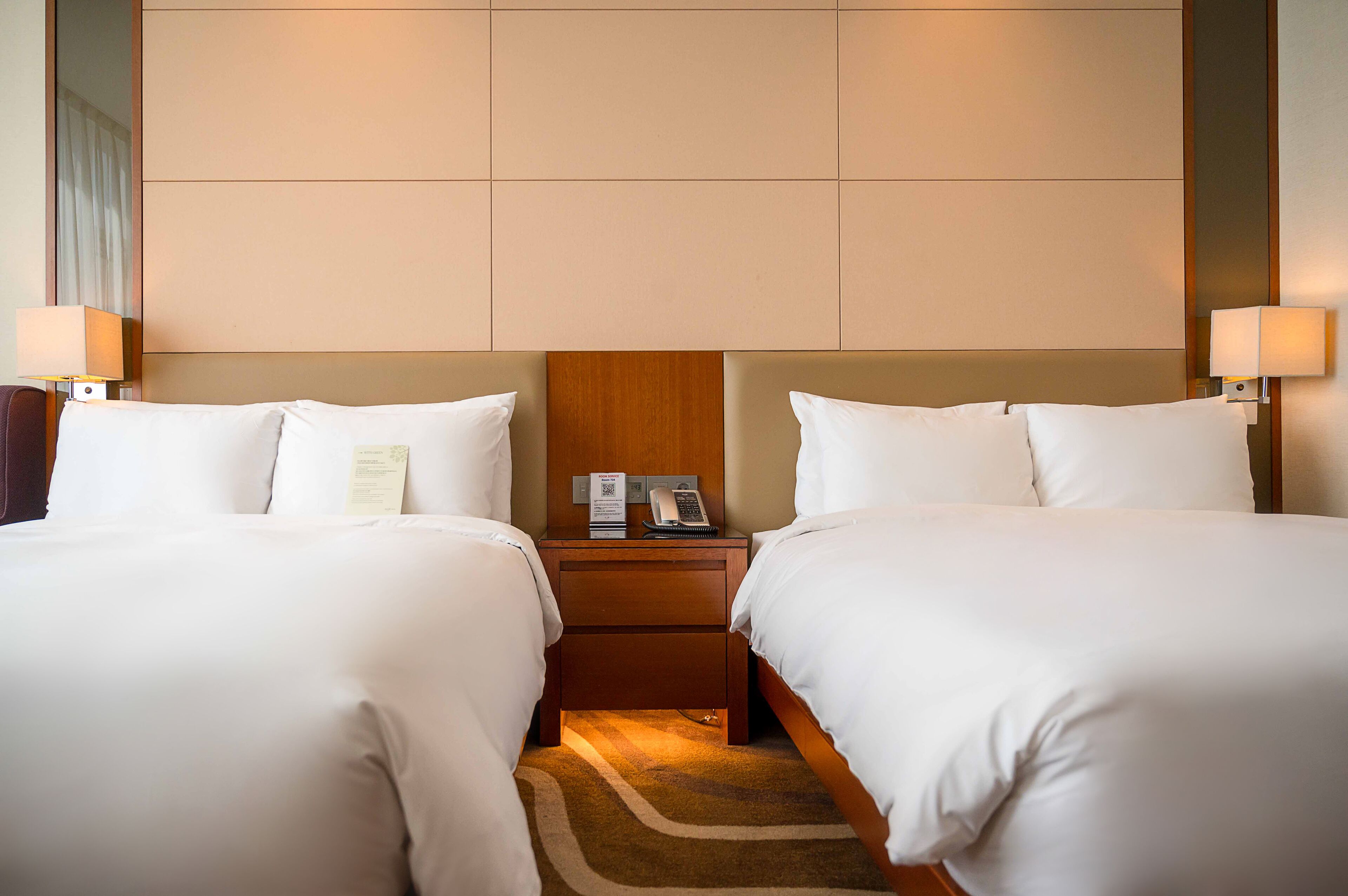 A hotel room with two beds, a wooden headboard, bedside lamps, a nightstand with a phone and a menu, and a carpeted floor.