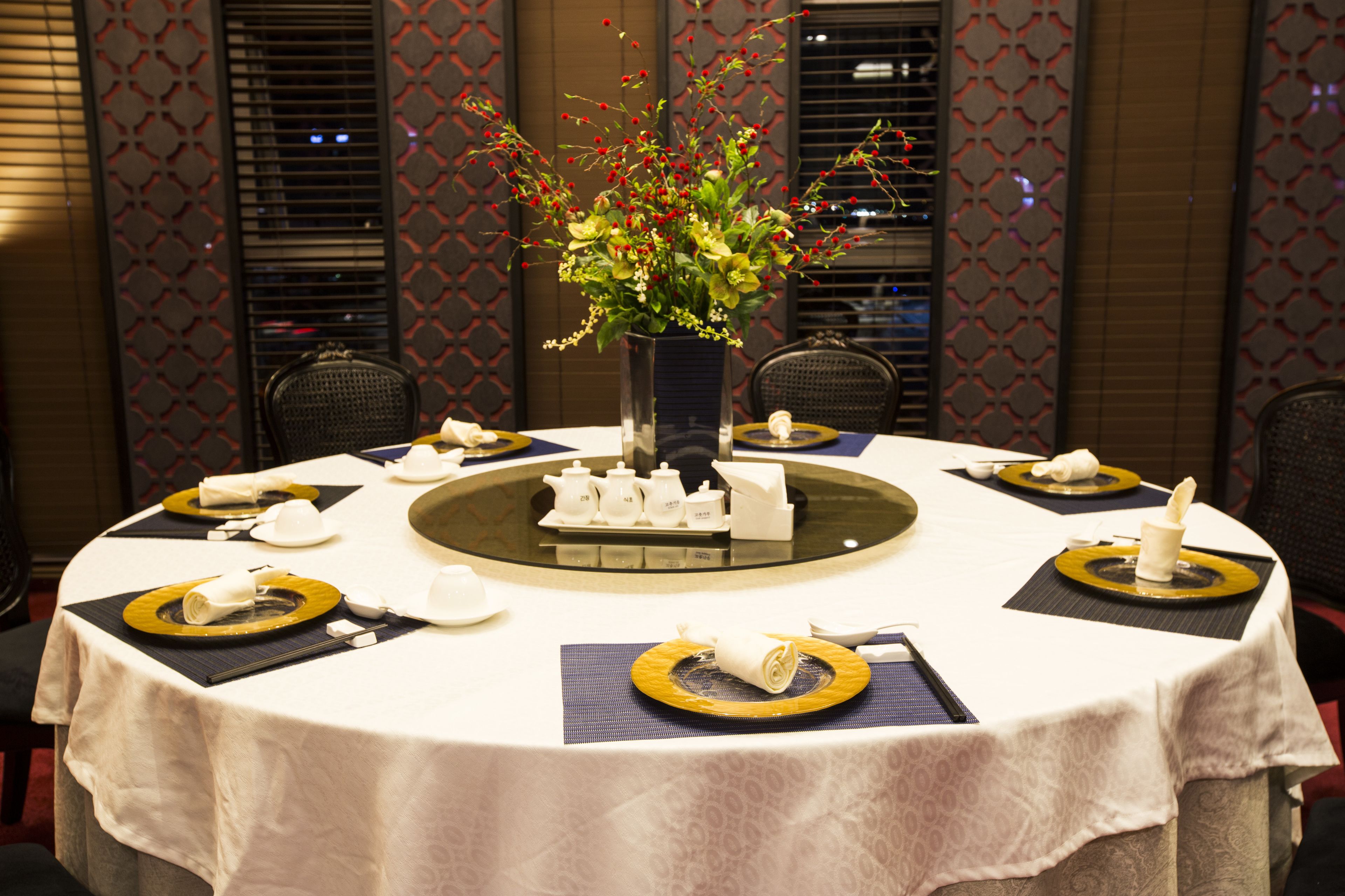 Een ronde eettafel, gedekt met witte tafelkleden, gouden en zwarte placemats en een bloemenarrangement.