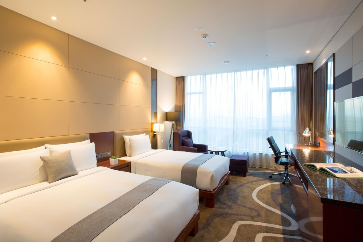 A hotel room with two beds, a desk, a chair, and a large window with curtains.