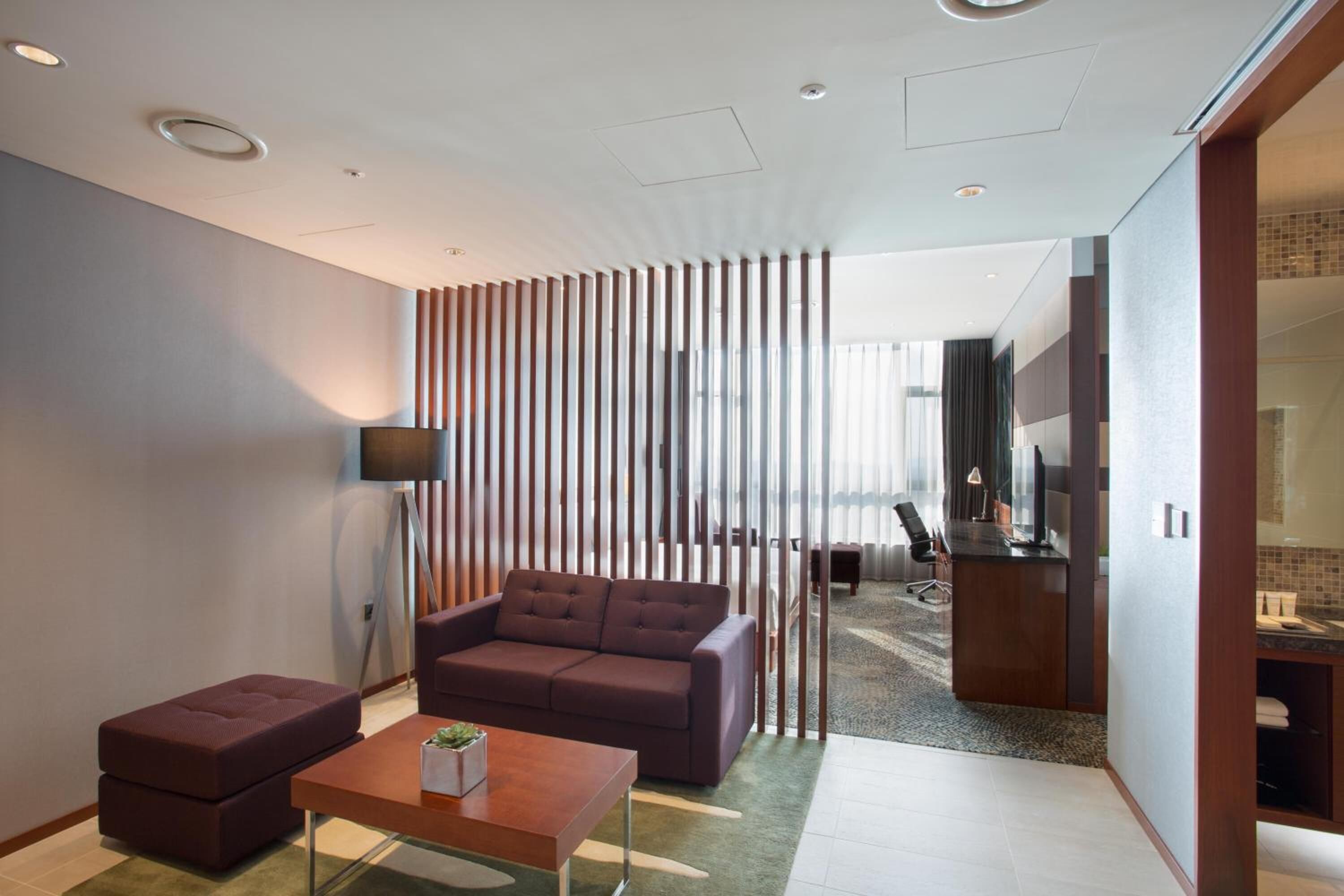 Una sala de estar moderna con sofá, reposapiés y mesa de centro. Hay un televisor sobre un mueble bajo, un separador de madera y vistas a otra habitación con un escritorio y una silla.