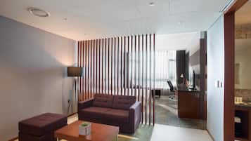 Una sala de estar moderna con sofá, reposapiés y mesa de centro. Hay un televisor sobre un mueble bajo, un separador de madera y vistas a otra habitación con un escritorio y una silla.