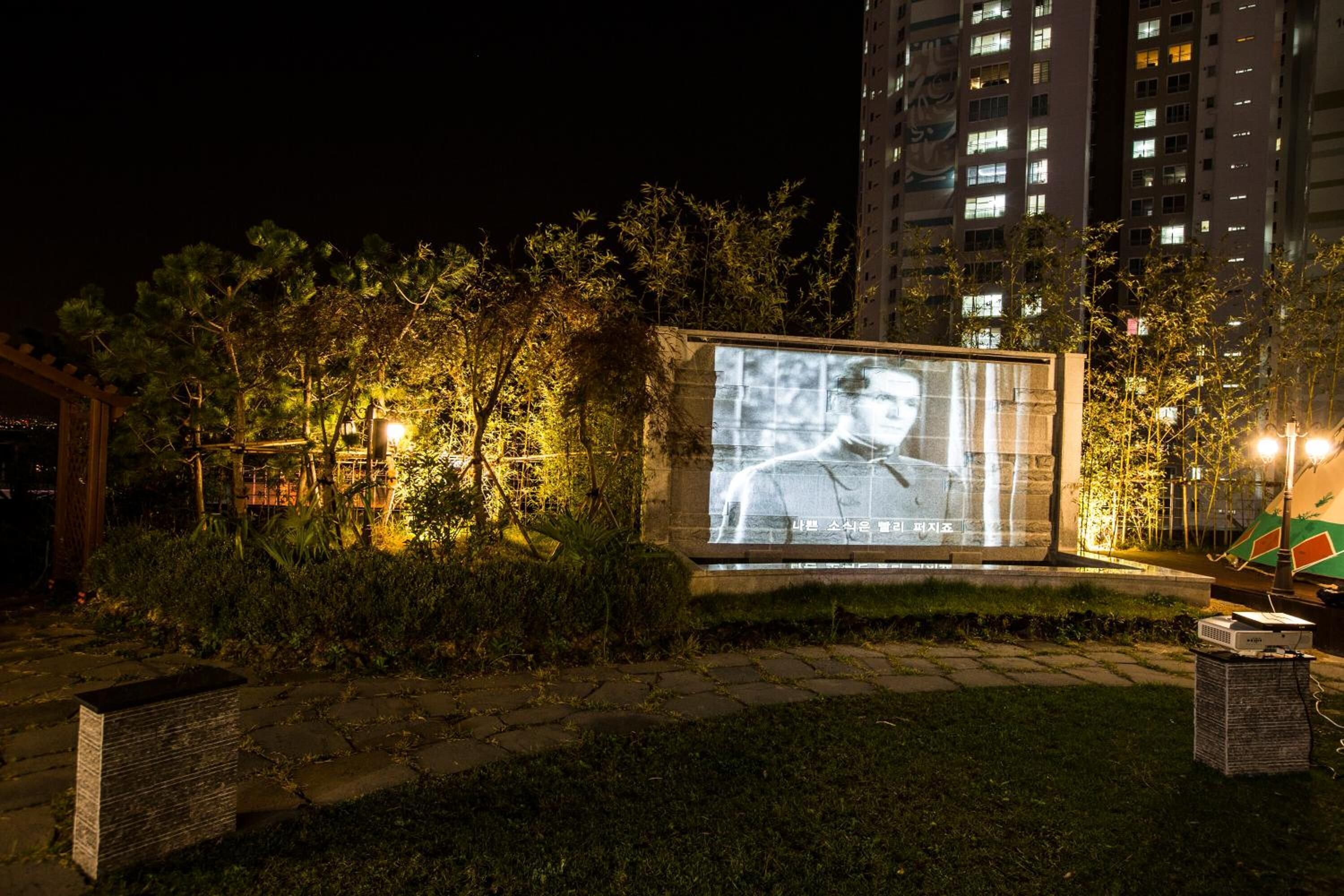 A nighttime outdoor area with a large illuminated billboard, a stone pathway, and a building in the background.