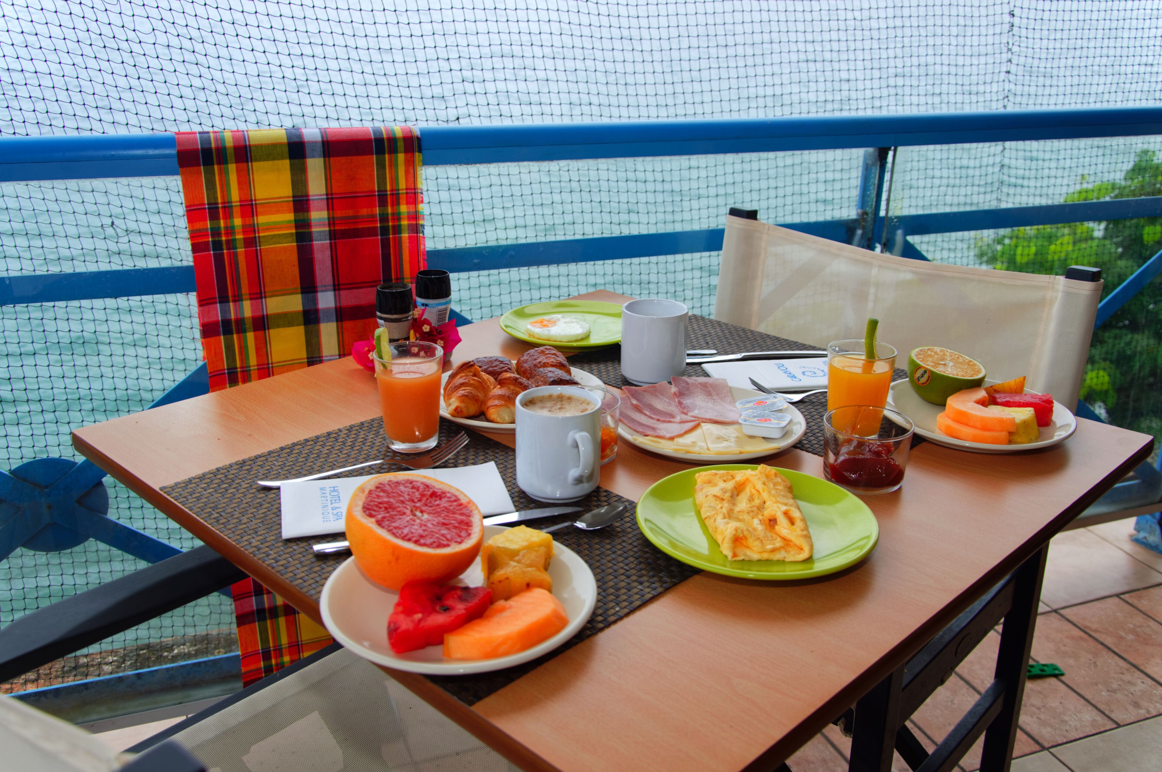 A breakfast table with various food items and drinks, set against a blue railing and netting.