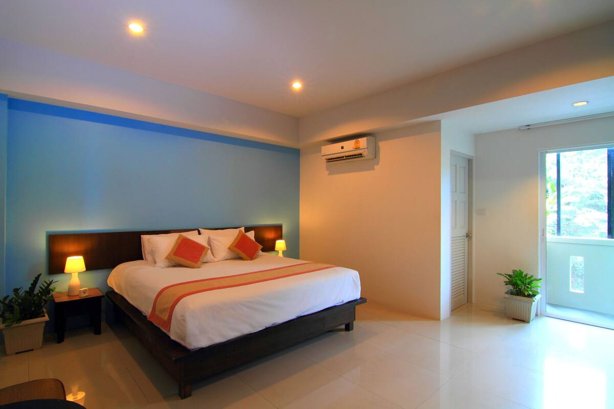 A hotel room with a large bed, bedside lamps, a potted plant, and an air conditioning unit.