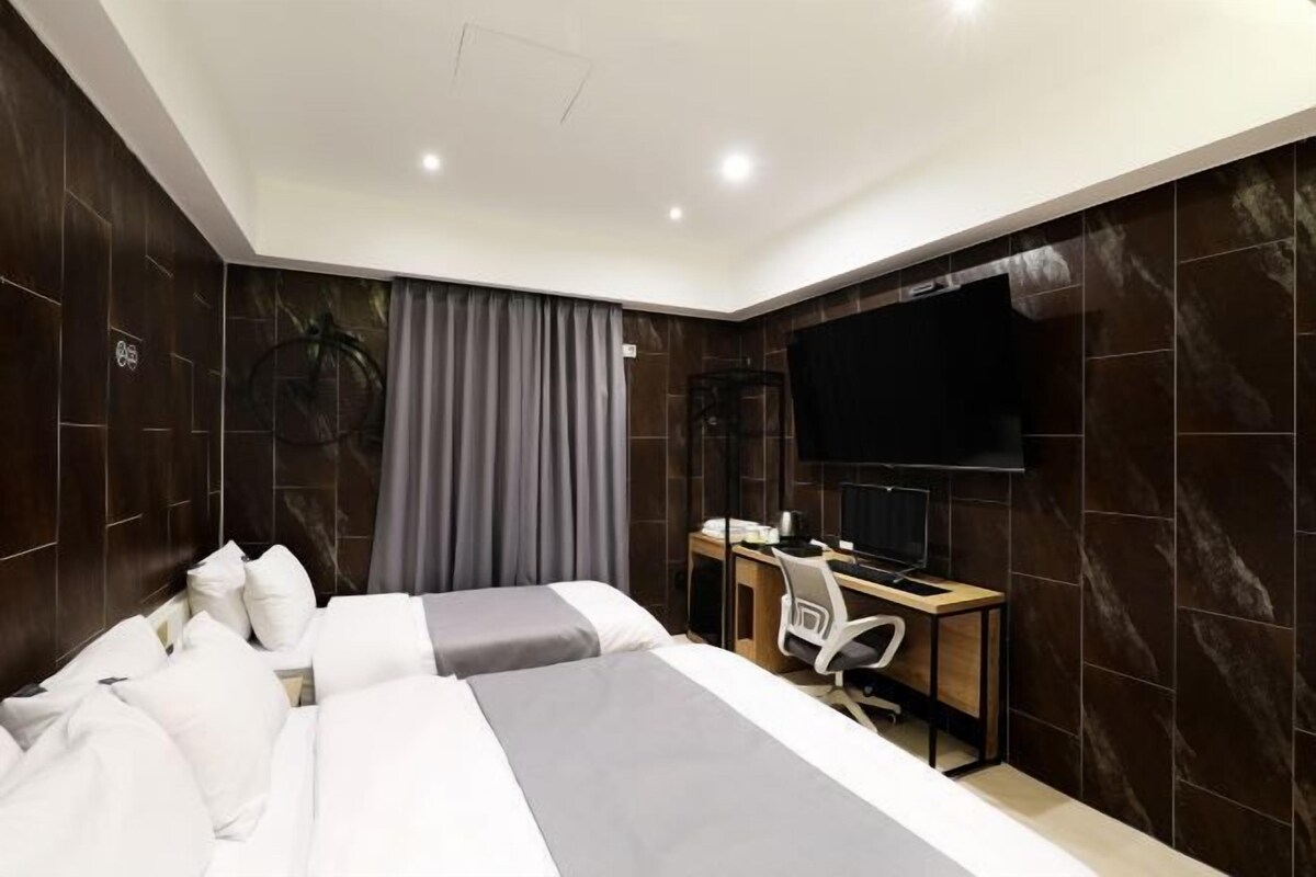 A modern hotel room with a large bed, a desk with a chair, a flat-screen TV, and a large window with curtains.