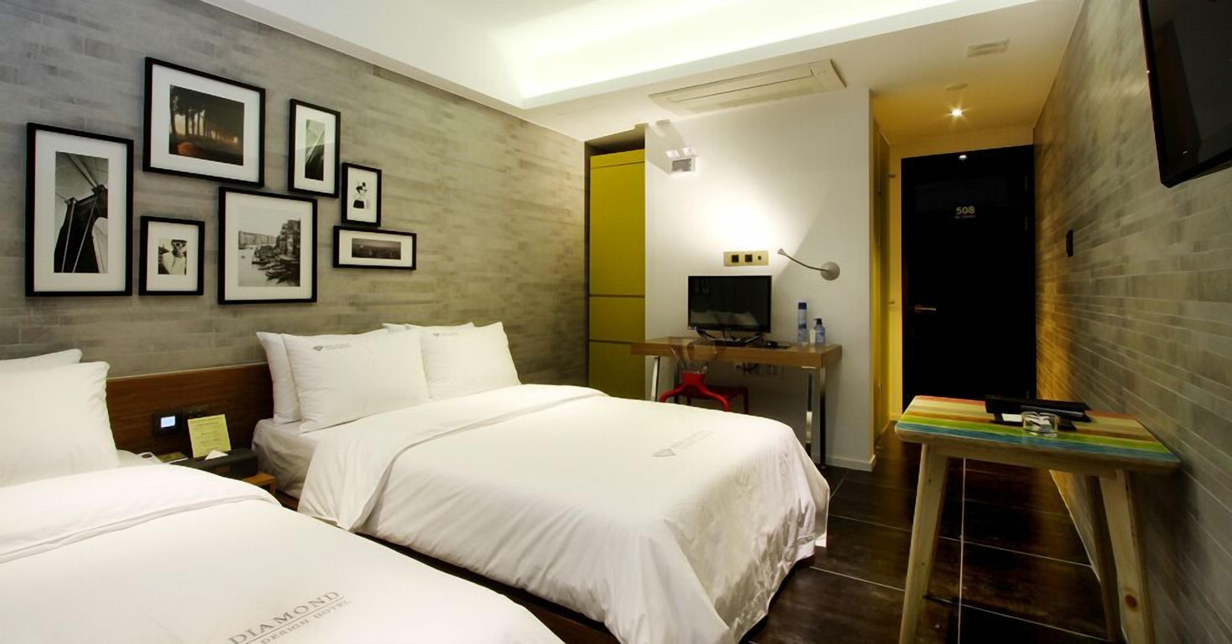 A hotel room with two beds, a desk with a computer, a TV, and framed pictures on the wall.