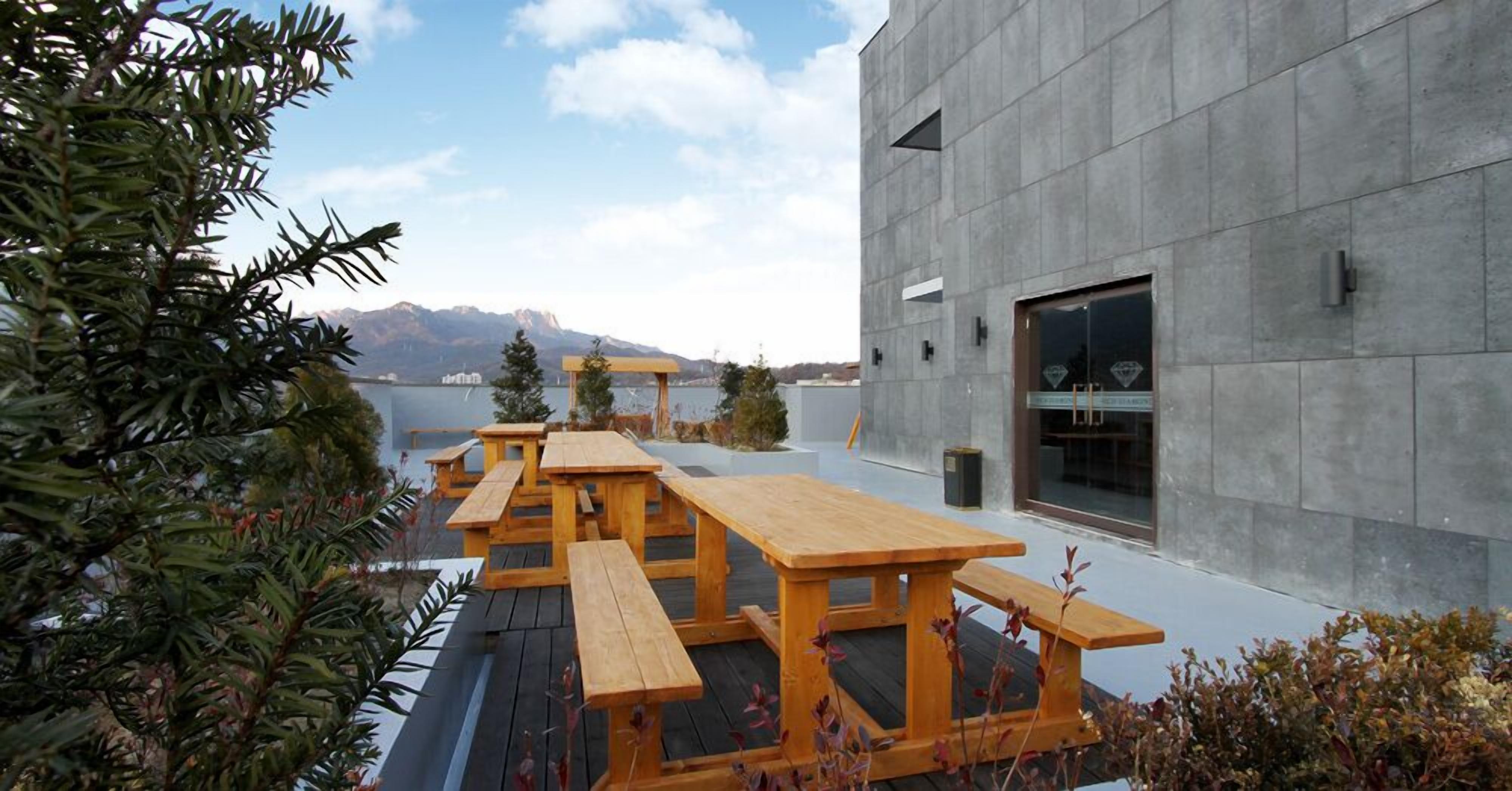 An outdoor seating area with wooden picnic tables, a modern building with a large window, and a view of mountains in the background.