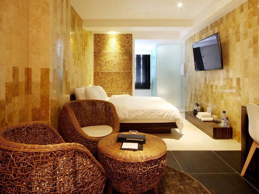 Una habitación de hotel moderna con una cama grande, dos sillones de mimbre, una mesita redonda pequeña, un televisor y un minibar.
