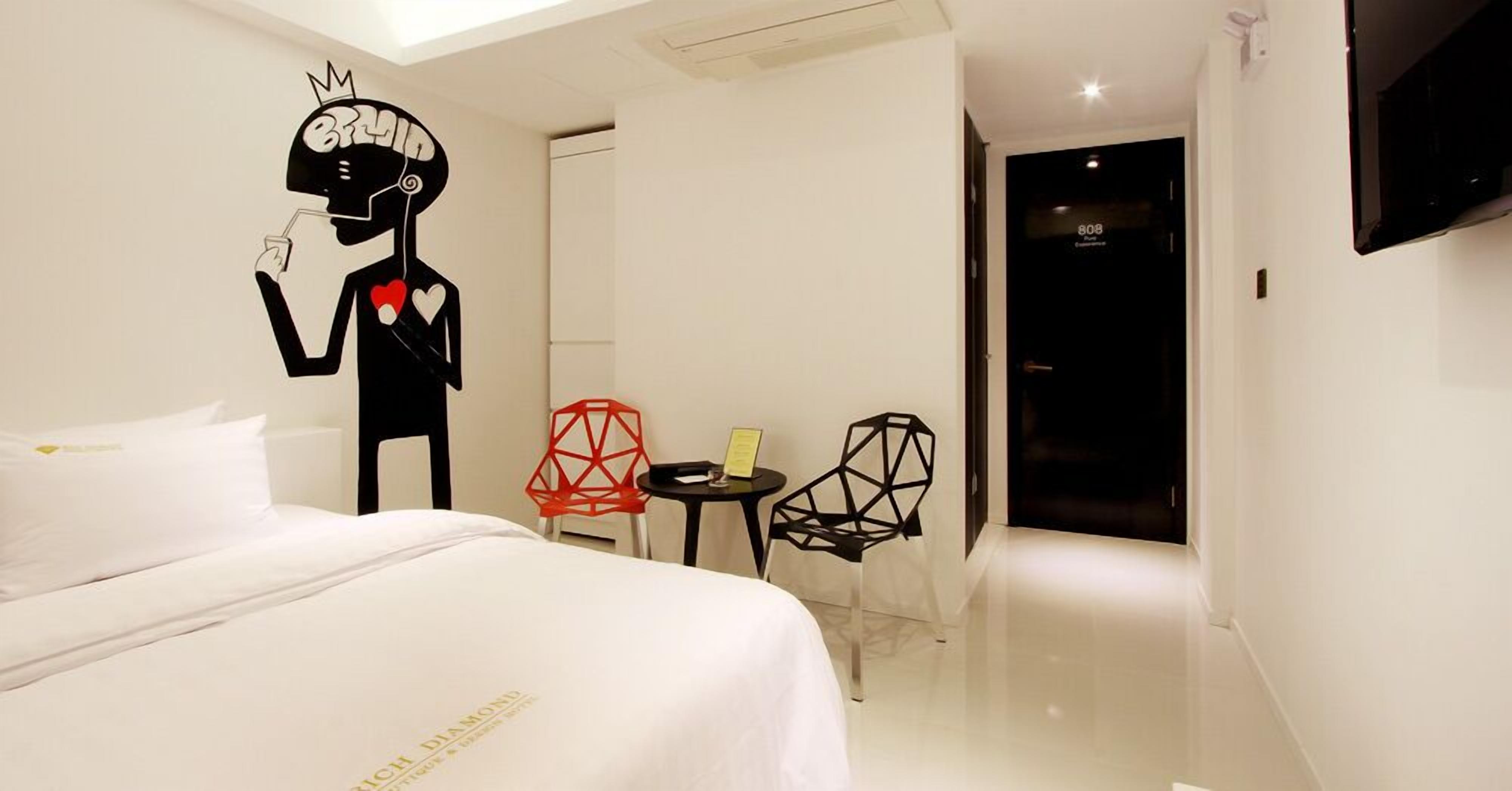 A modern hotel room with a bed, a wall-mounted TV, and geometric chairs.