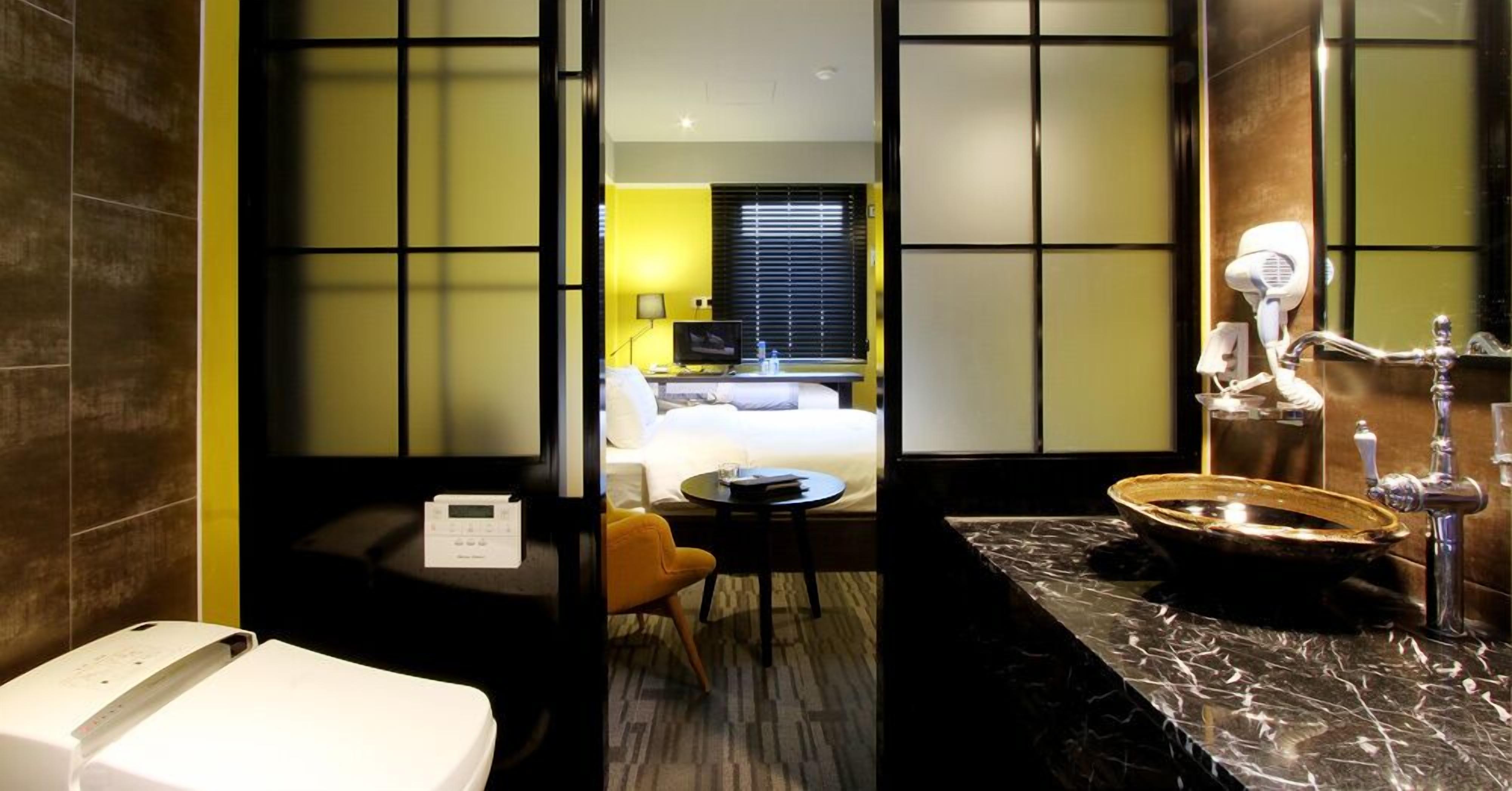 A hotel room with a bed, a desk, a chair, a bathroom with a sink and mirror, and a window with blinds.