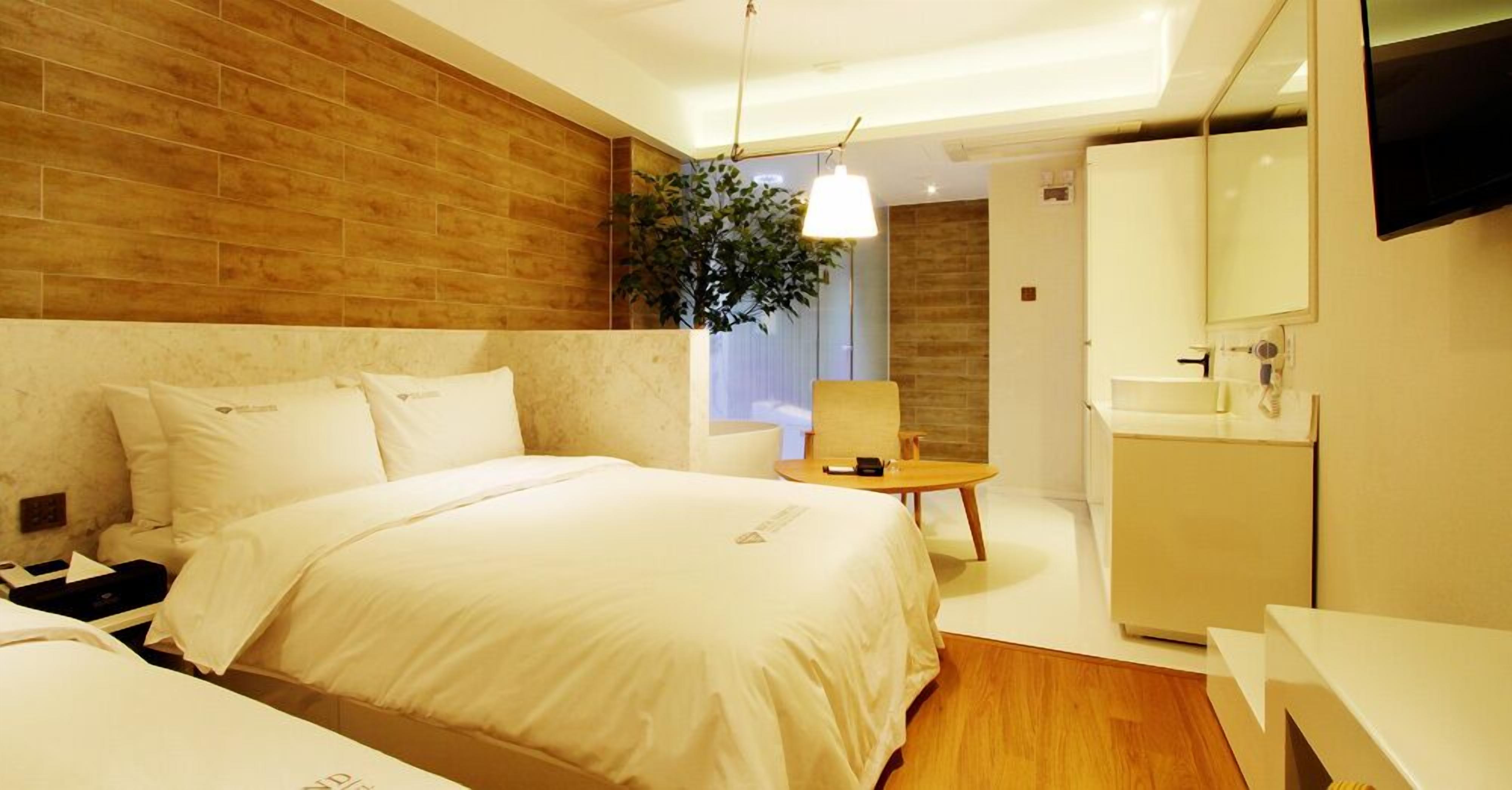 A modern hotel room with a large bed, a bedside table, a small desk, a chair, a plant, a mirror, and a bathroom with a sink and mirror.