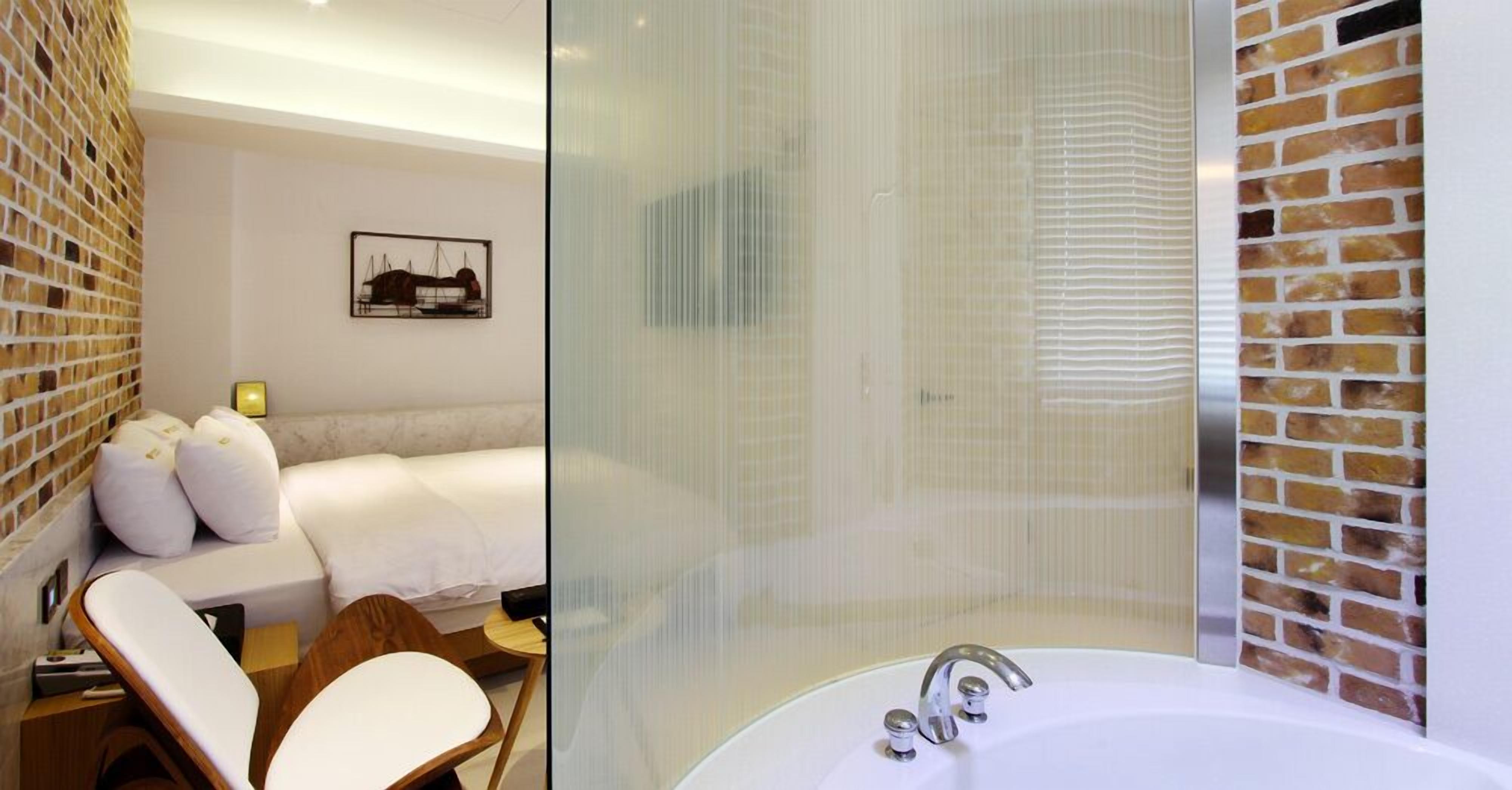 A modern hotel room with a large bed, a sitting area with a chair, a glass shower enclosure, and a brick wall design.