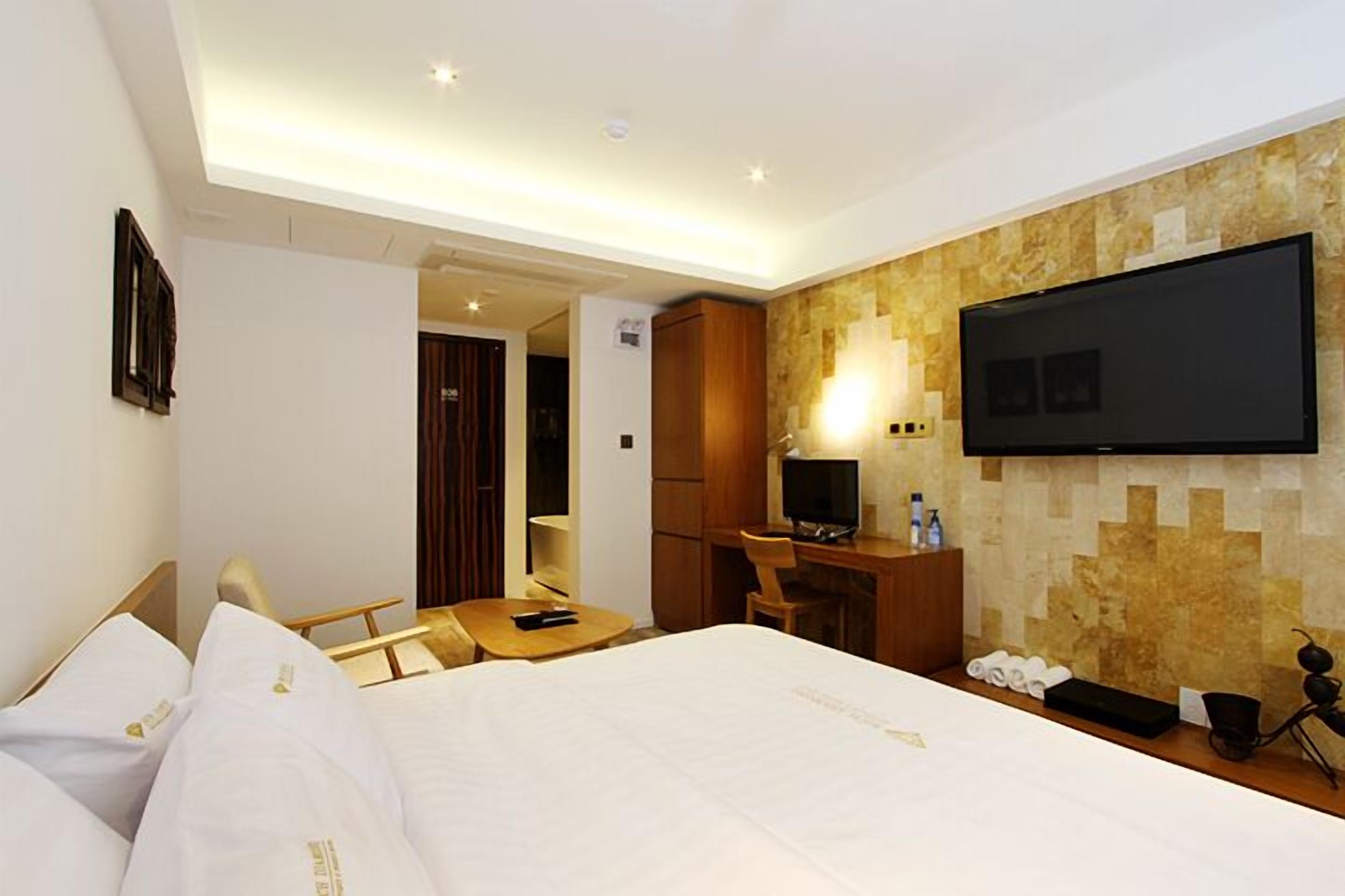 Habitación de hotel con cama, escritorio, silla, televisión y baño visible a través de una puerta abierta.