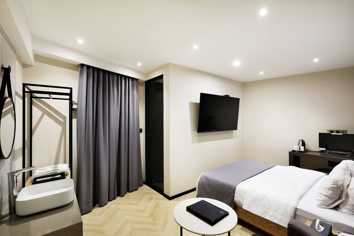A modern hotel room with a bed, a flat-screen TV, a small table, a bathroom with a sink and mirror, and a shower area.