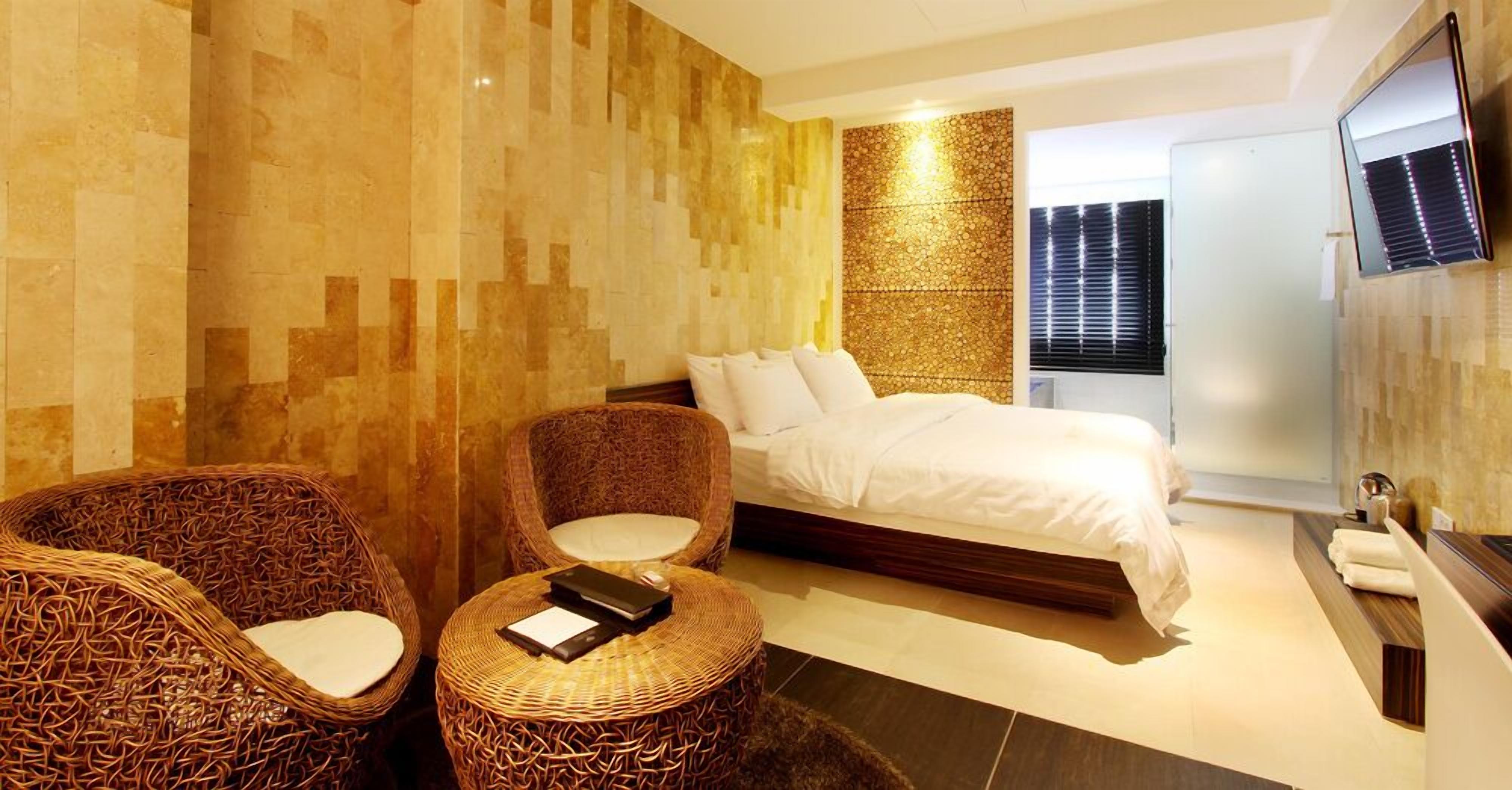 Une chambre d’hôtel moderne avec un grand lit, deux fauteuils en osier, une petite table ronde et un téléviseur fixé au mur.