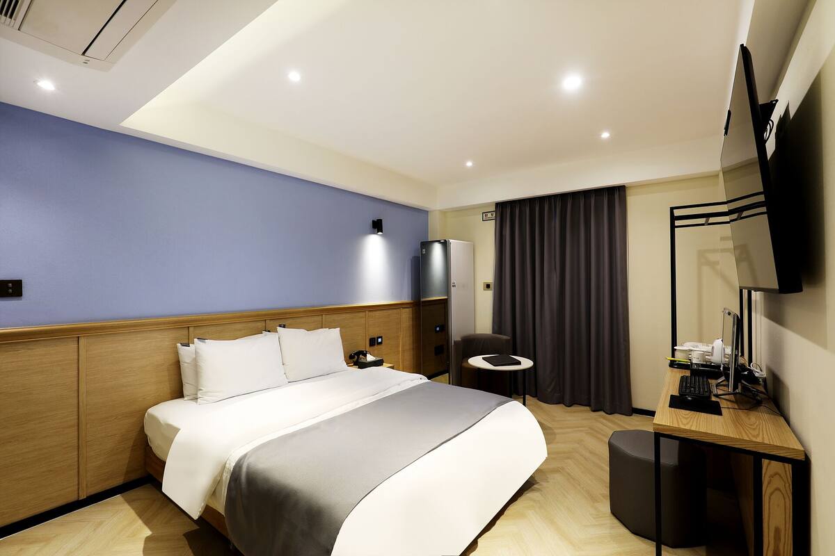 A modern hotel room with a bed, a desk, a chair, a television, and a large window with curtains.